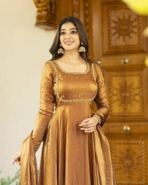 A stunning honey-gold Anarkali dress crafted in rich Chinnon silk fabric, featuring a graceful flared silhouette. The bodice is adorned with delicate golden embroidery and tiny floral motifs, complemented by a square neckline and full-length sleeves for an elegant finish. The subtle sheen of the fabric adds a regal charm, making it perfect for festive occasions, weddings, and traditional celebrations. Paired with statement earrings, this outfit exudes timeless sophistication. ✨